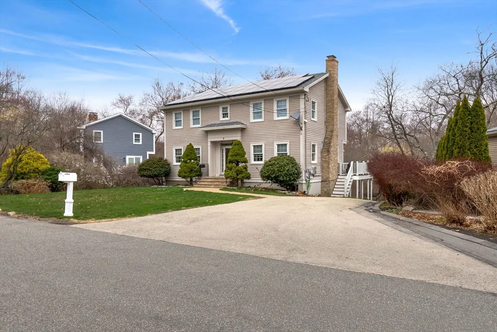 Beige two-story house with solar panels, chimney, and white trim on a sunny day. A gravel driveway leads to the house.