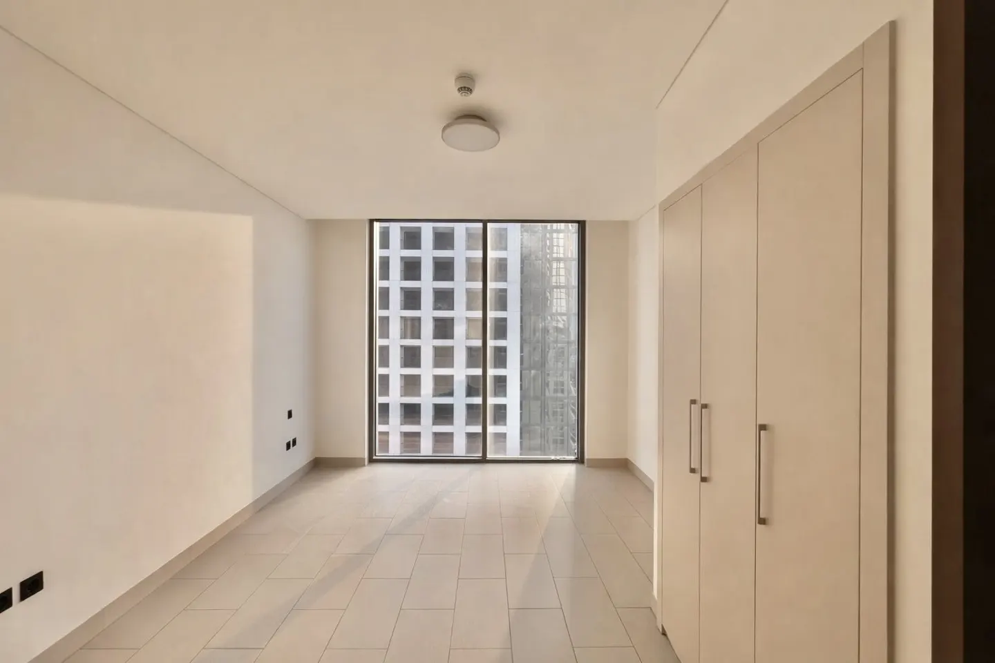 Empty room with beige tile floor, white walls, and a large window showing a city view. A light fixture is on the ceiling and a wardrobe is on the right.
