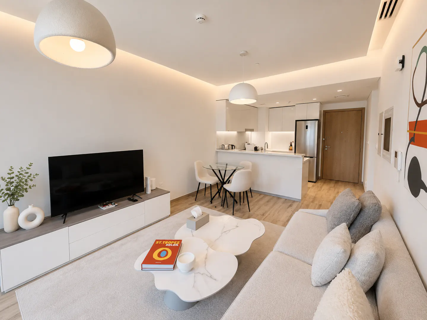 Bright, modern apartment interior with a white sofa, TV, and kitchen. A unique coffee table sits on a light rug.