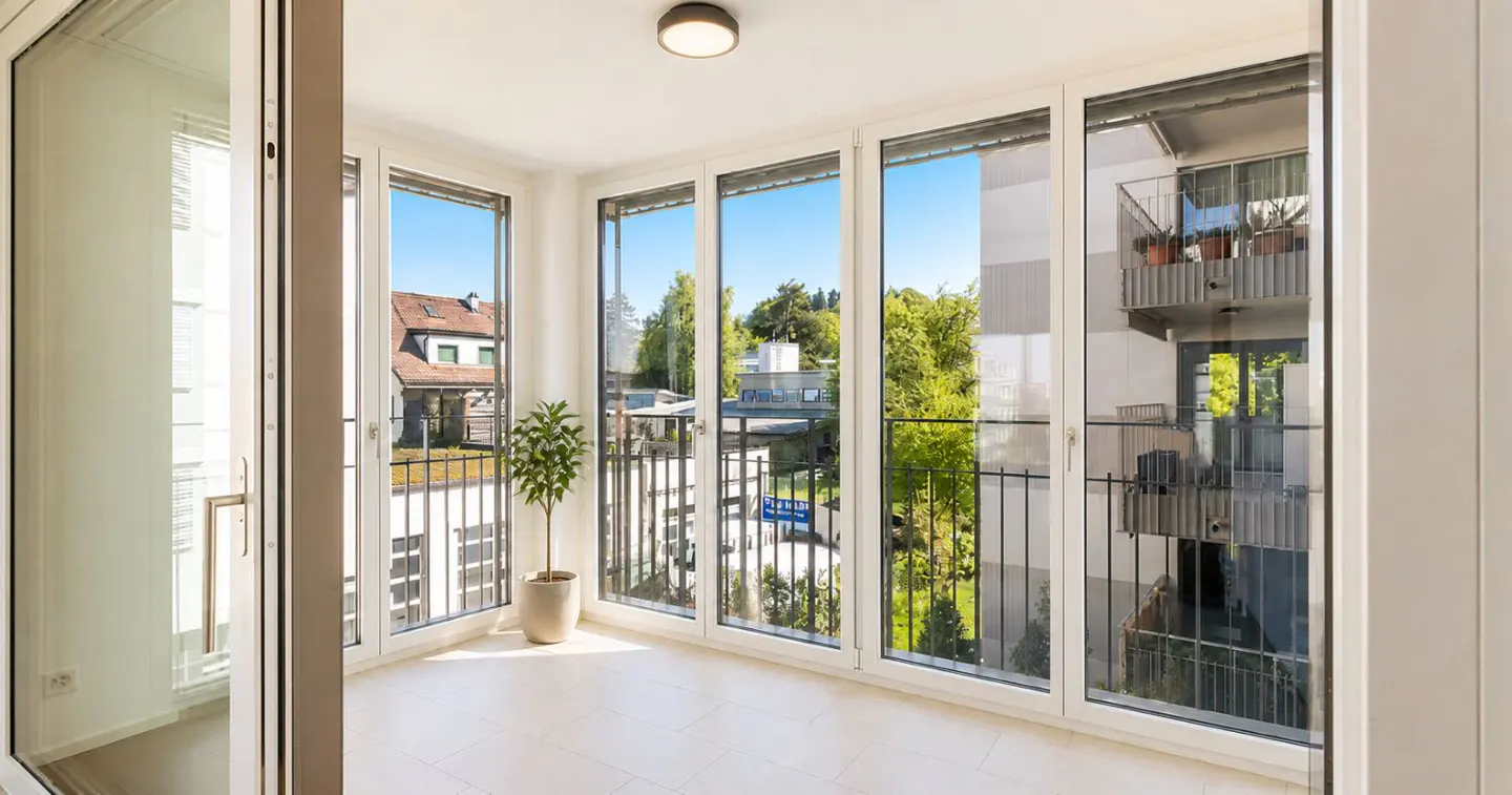 Bright sunroom with white tile floor and large windows. A potted plant sits in the corner, and a view of trees and buildings is visible outside.