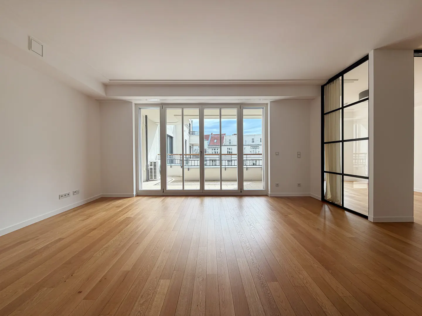 Bright, empty room with light wood floors, white walls, and a large glass door leading to a balcony with city views. Black framed glass wall on the right.