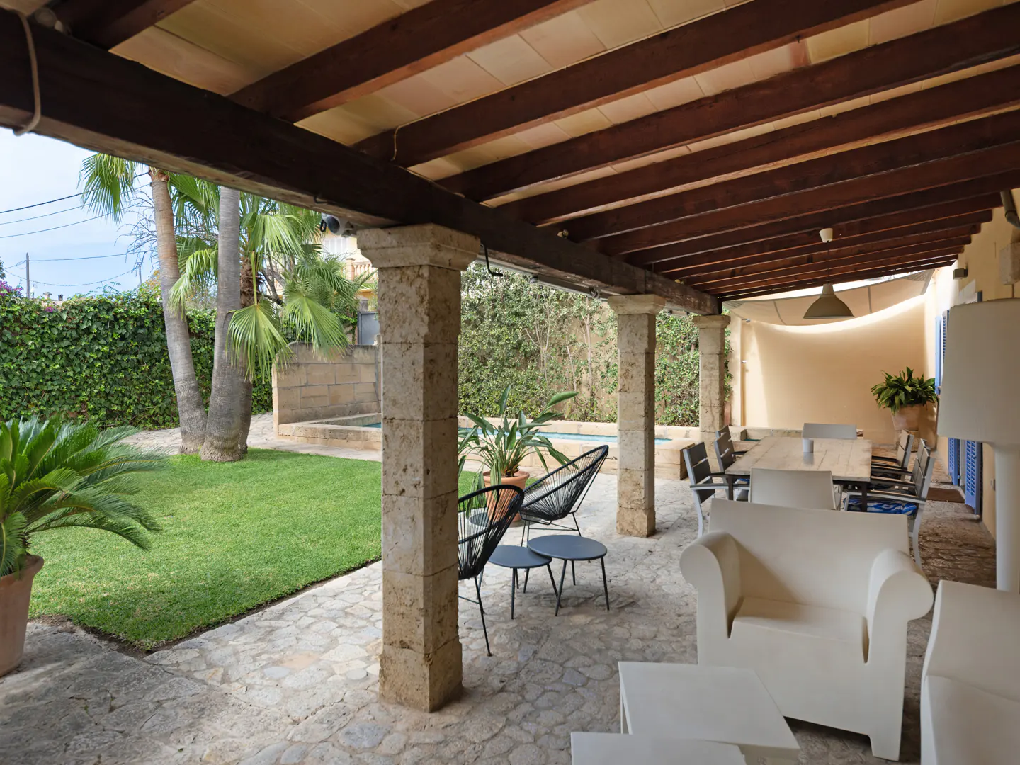 Covered patio with stone pillars, dark wood beams, and stone floor. White modern furniture and a dining table are visible. Green lawn and pool in the background.
