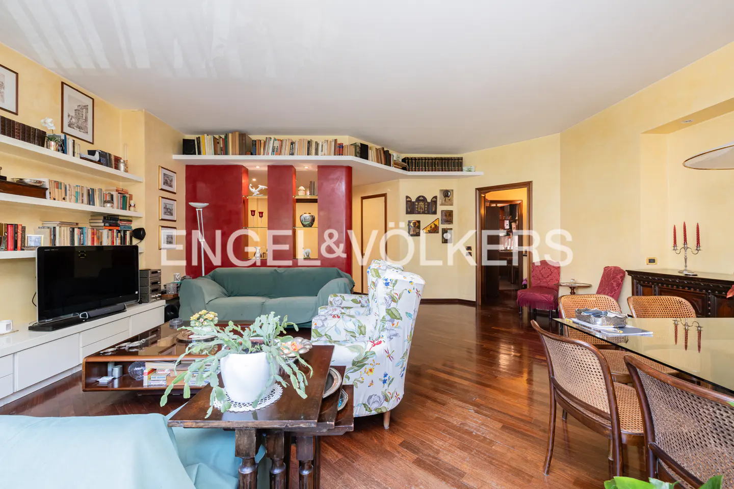Living room with yellow walls, hardwood floors, a green sofa, floral armchair, and a dining area with rattan chairs.