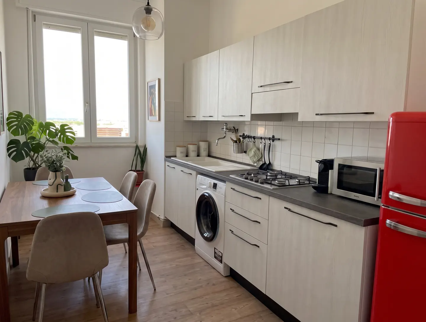 Bright kitchen with a wooden table, beige chairs, white cabinets, a red fridge, and a washing machine. A window is on the left.