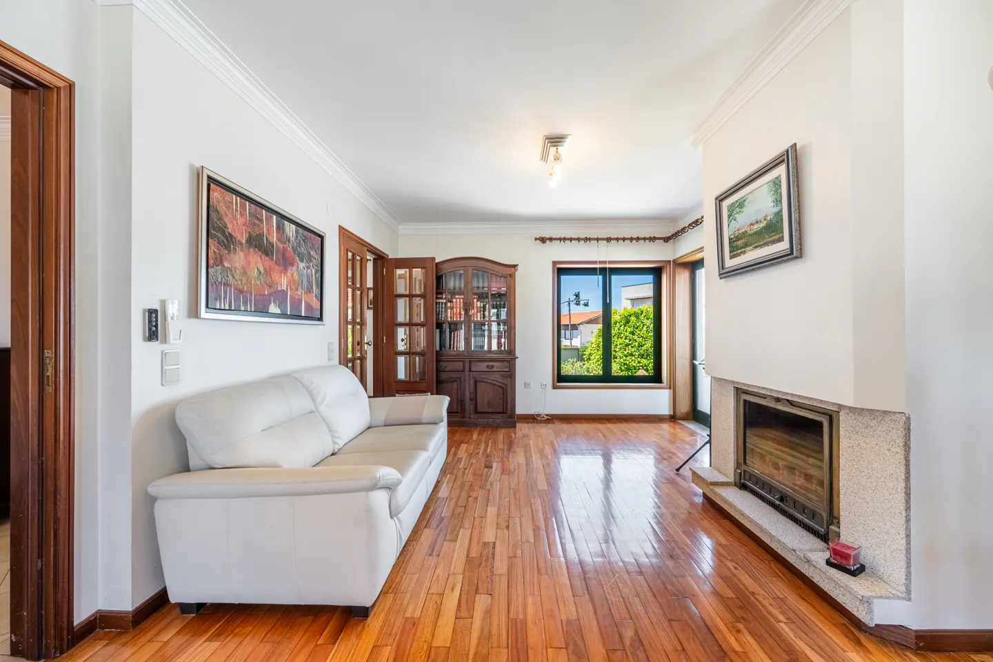 Bright living room with hardwood floors, white sofa, fireplace, and a dark wood bookcase. A window shows a green yard.