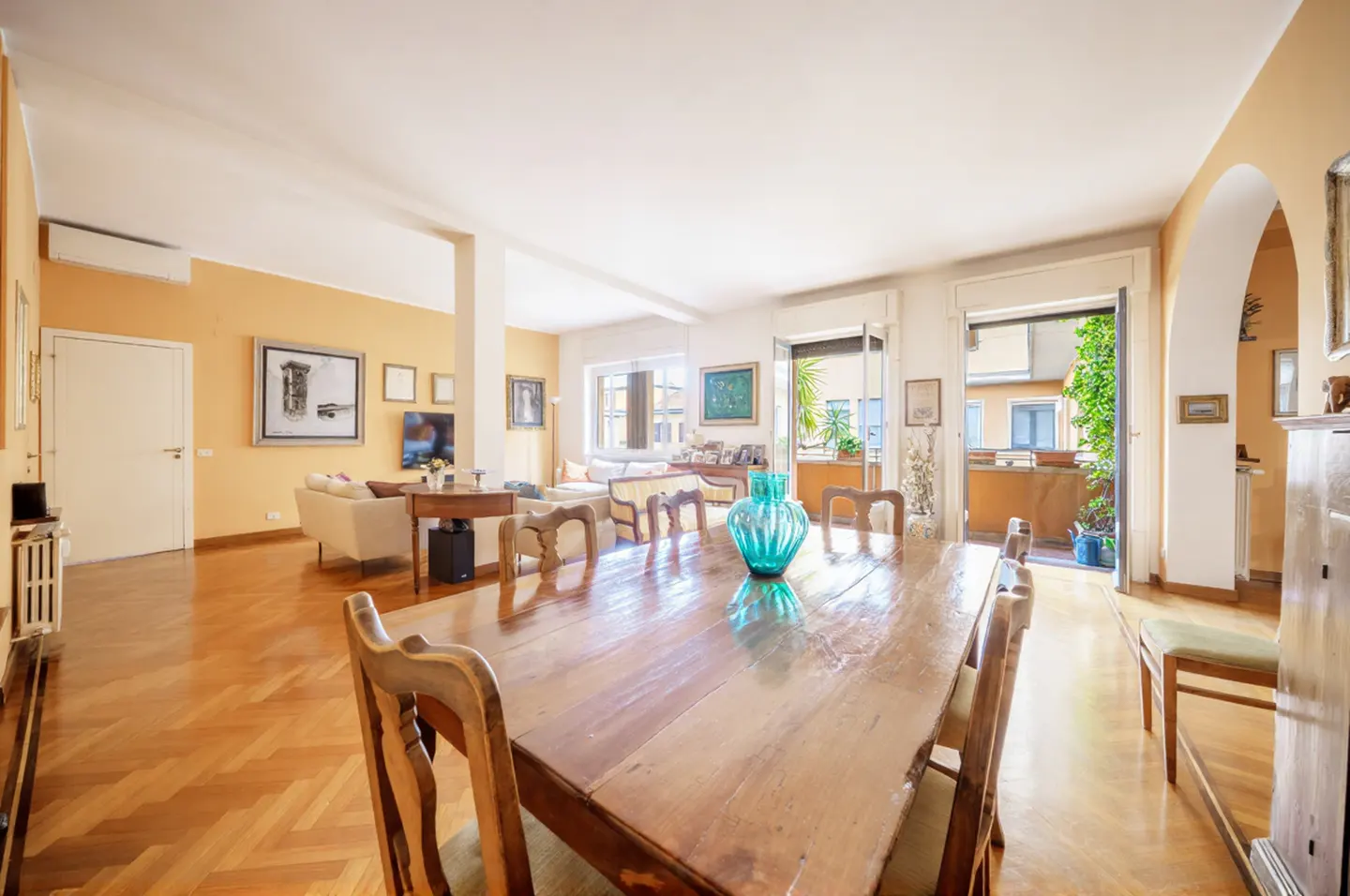 A bright, open-concept living and dining area with a long wooden table, chairs, and a turquoise vase. Balcony doors open to a view of greenery.