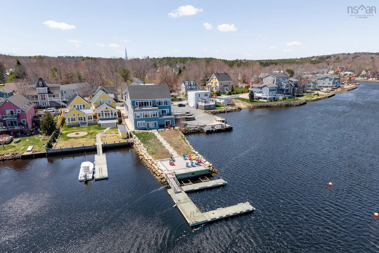 Aerial view of waterfront homes with docks on a sunny day. Blue house with balconies, colorful chairs, and boats visible.