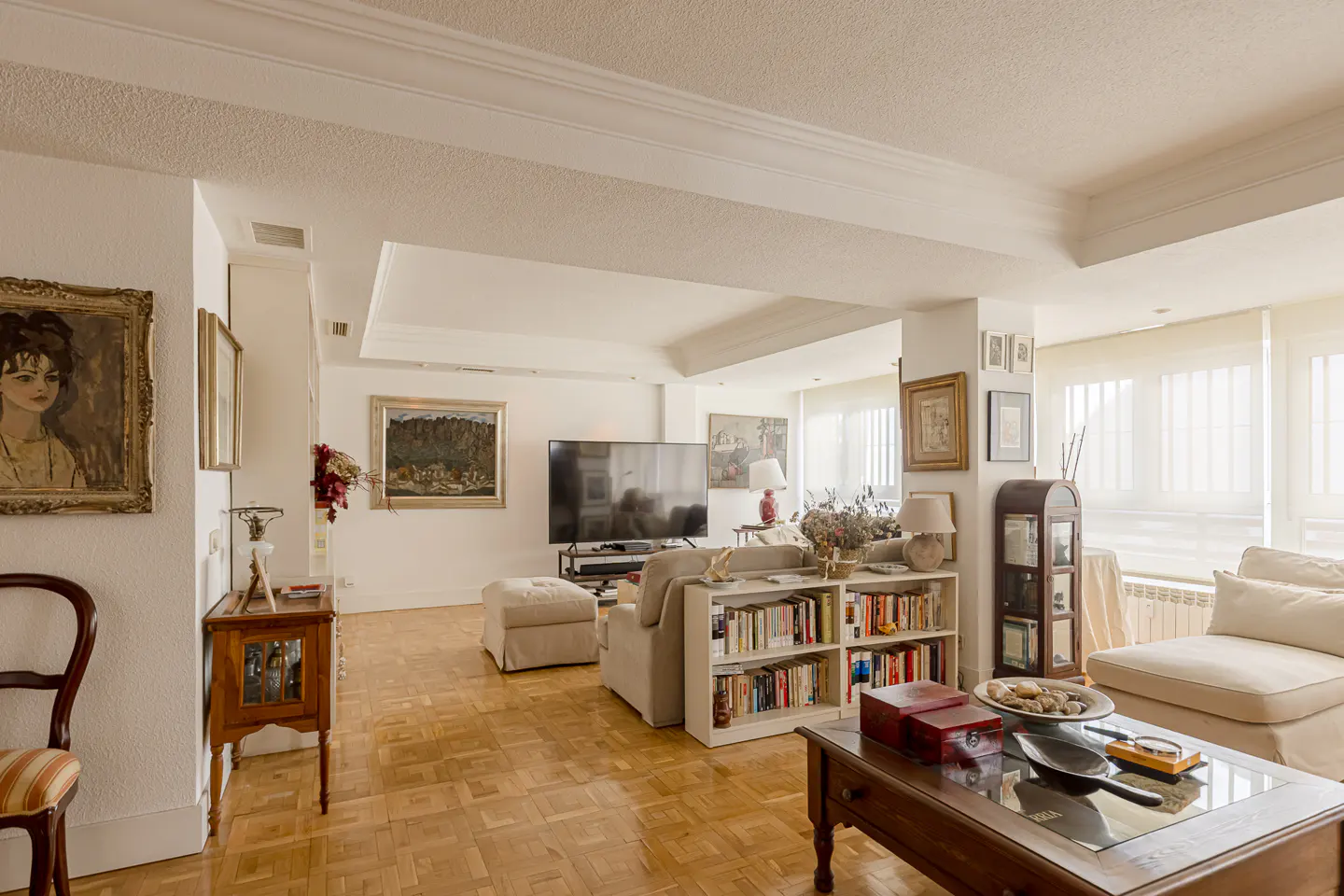 Bright living room with parquet floors, beige sofa, bookshelf, large TV, and framed art on white walls.