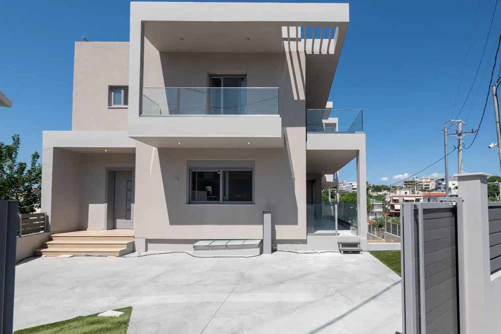 Modern beige two-story house with a concrete driveway, glass-railed balconies, and a clear blue sky background.