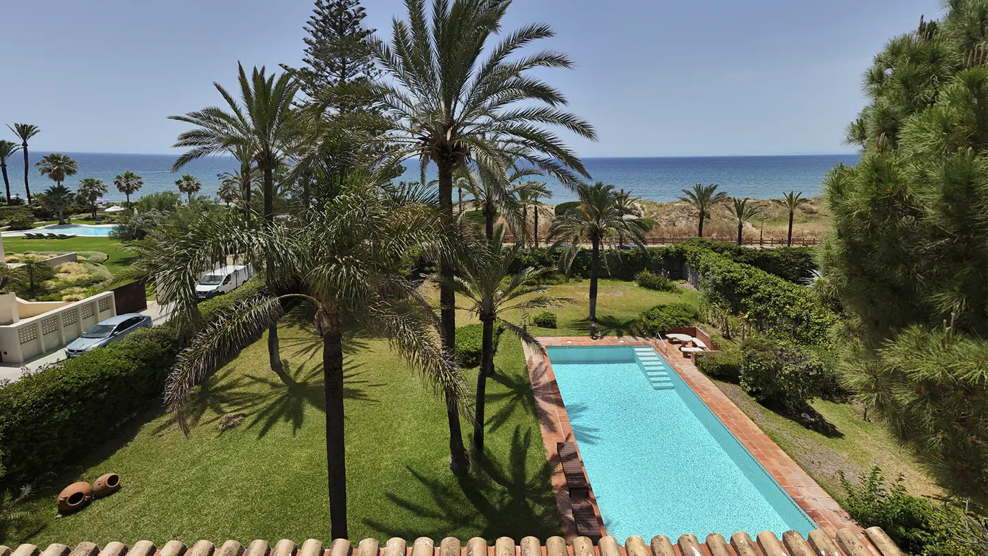 View of a turquoise pool surrounded by green grass and palm trees, with the ocean visible in the background.