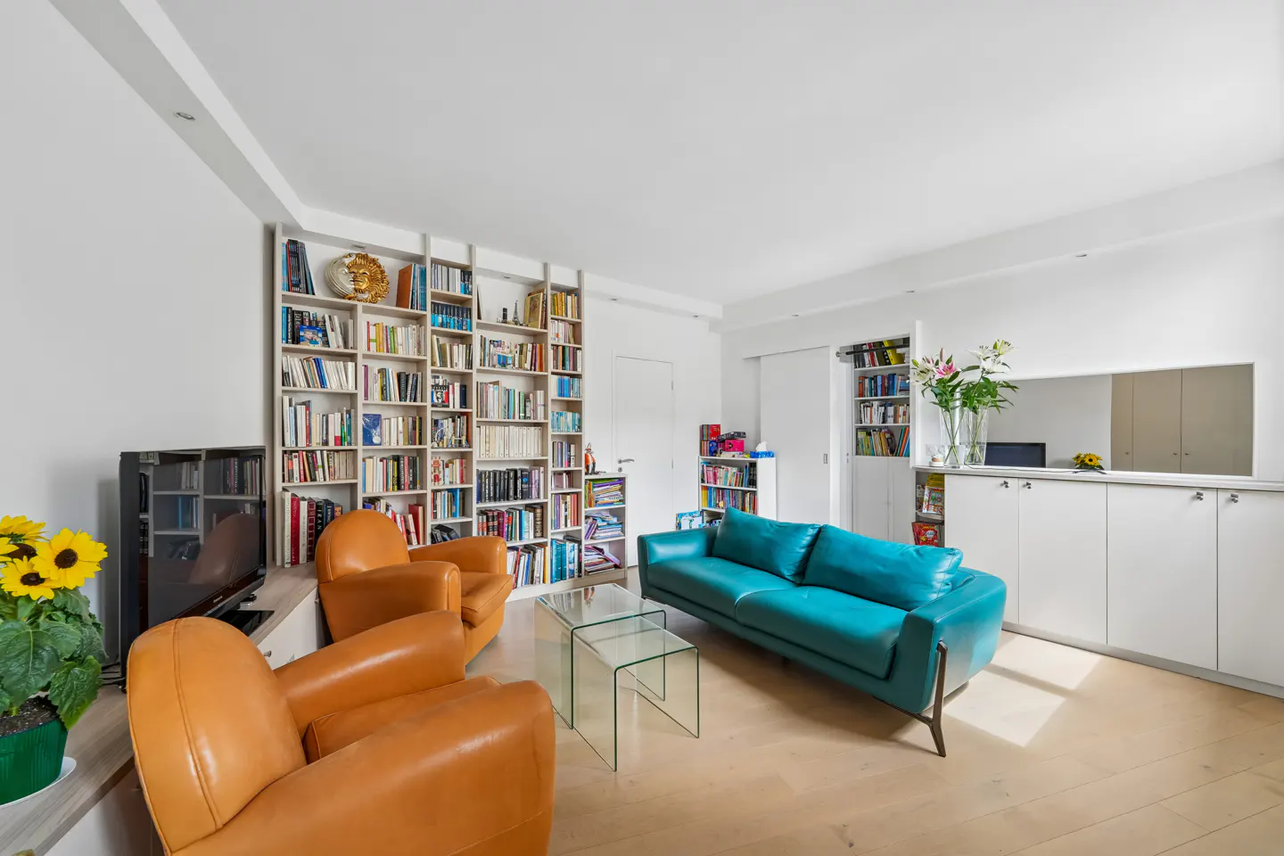 Bright living room with a turquoise sofa, two orange armchairs, glass tables, and a large bookshelf filled with books.