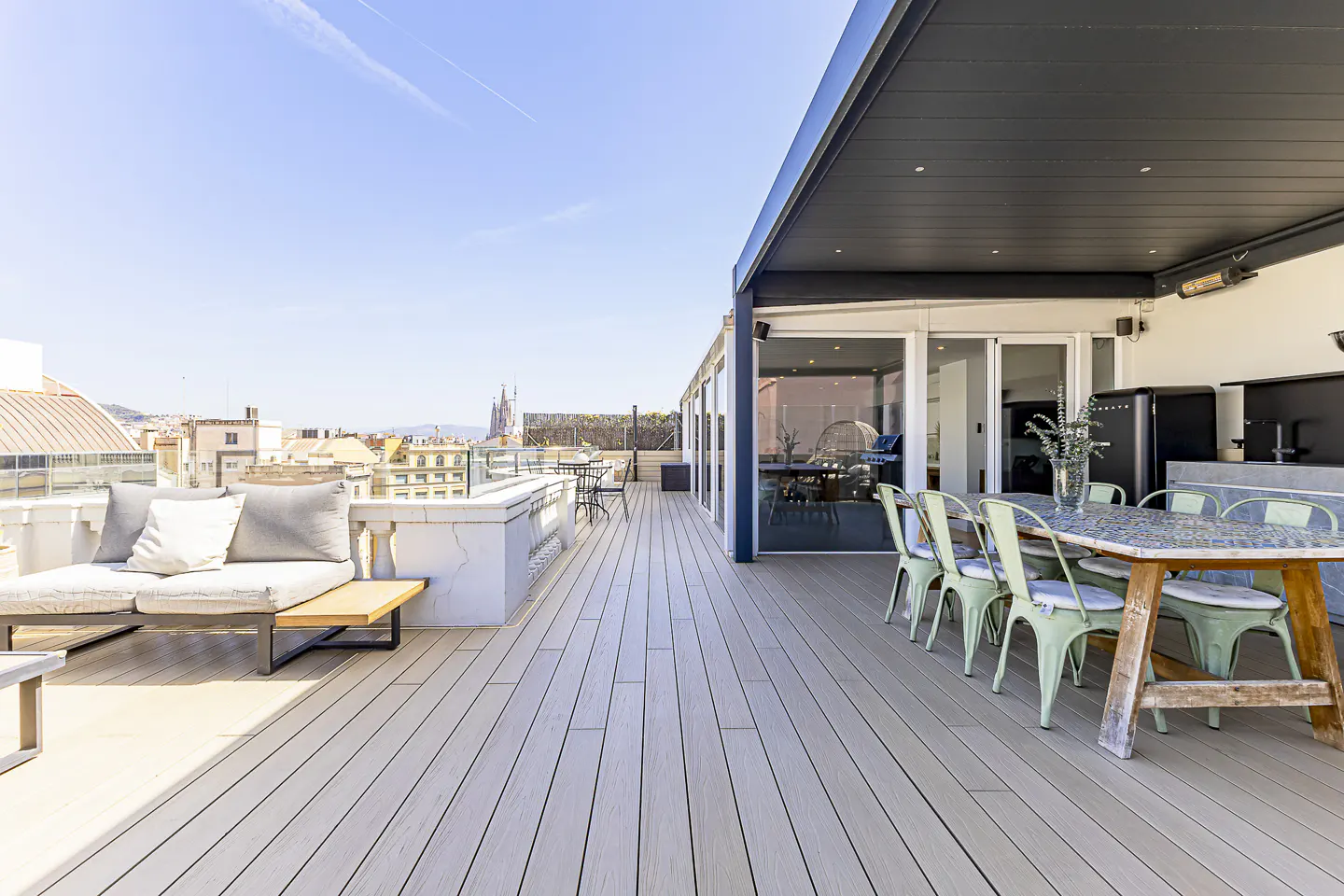 A rooftop patio with a dining table, chairs, and a lounge area, overlooking a cityscape under a blue sky.