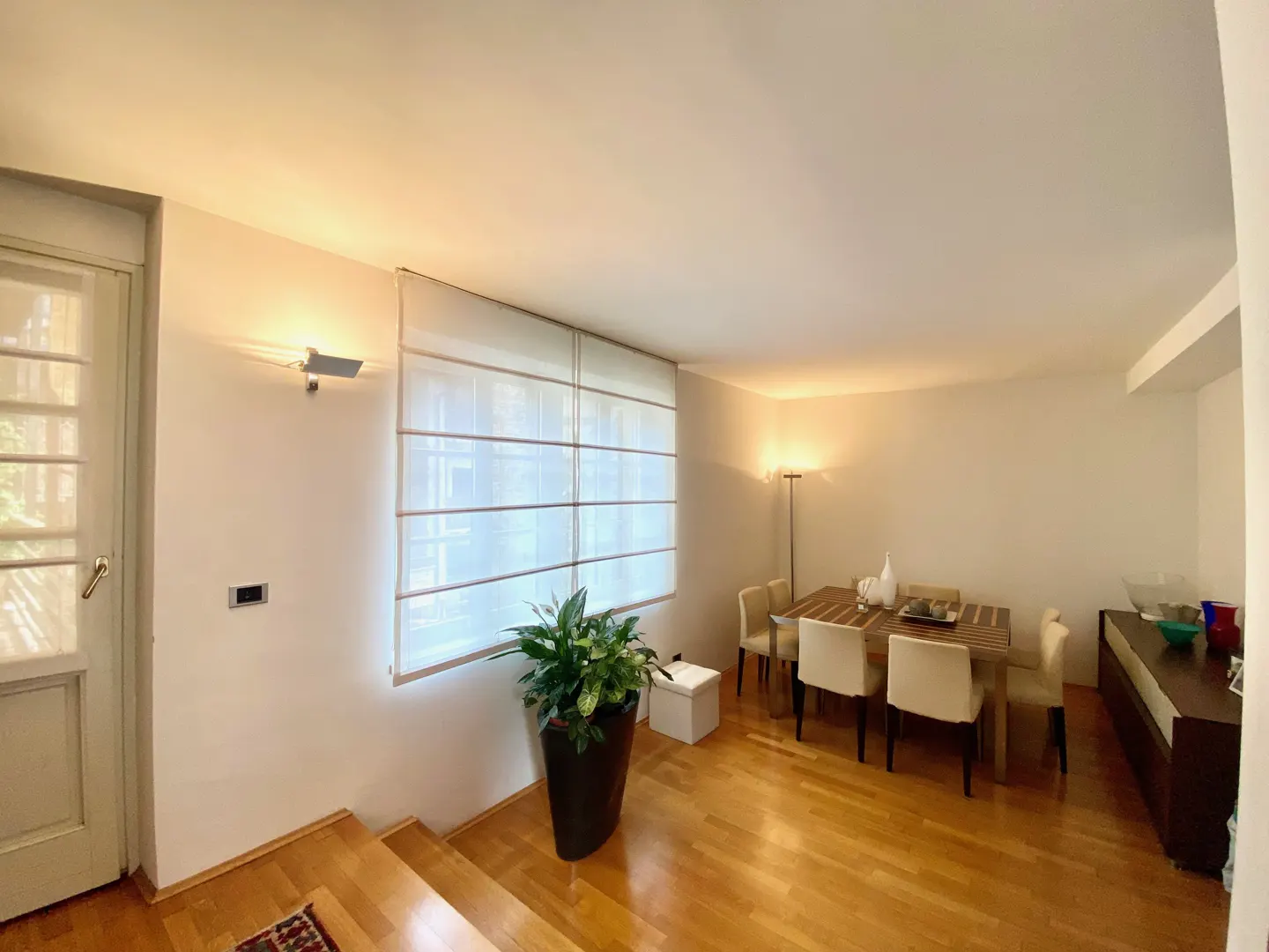 Bright dining room with hardwood floors, a wooden table with six chairs, and a large potted plant near a window with white blinds.