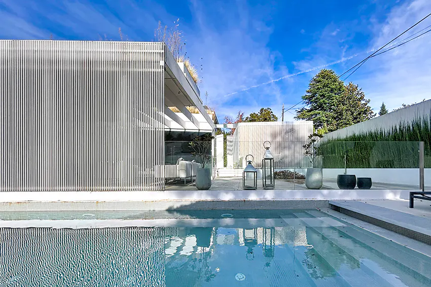Modern home exterior with a pool. White vertical slats cover part of the house. Lanterns and potted plants decorate the patio. Blue sky.