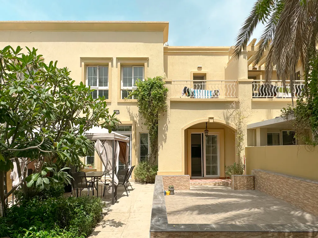 Backyard view of a two-story, light yellow house with a patio, table, chairs, and green trees.