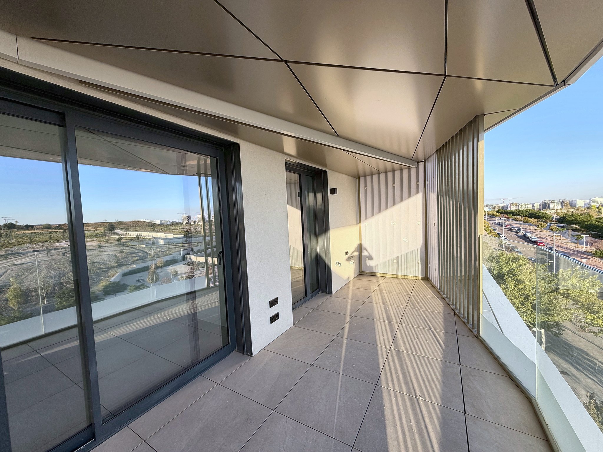 Modern apartment balcony with tiled floor, glass railings, and sliding doors overlooking a sunny urban landscape. [AI-generated text]