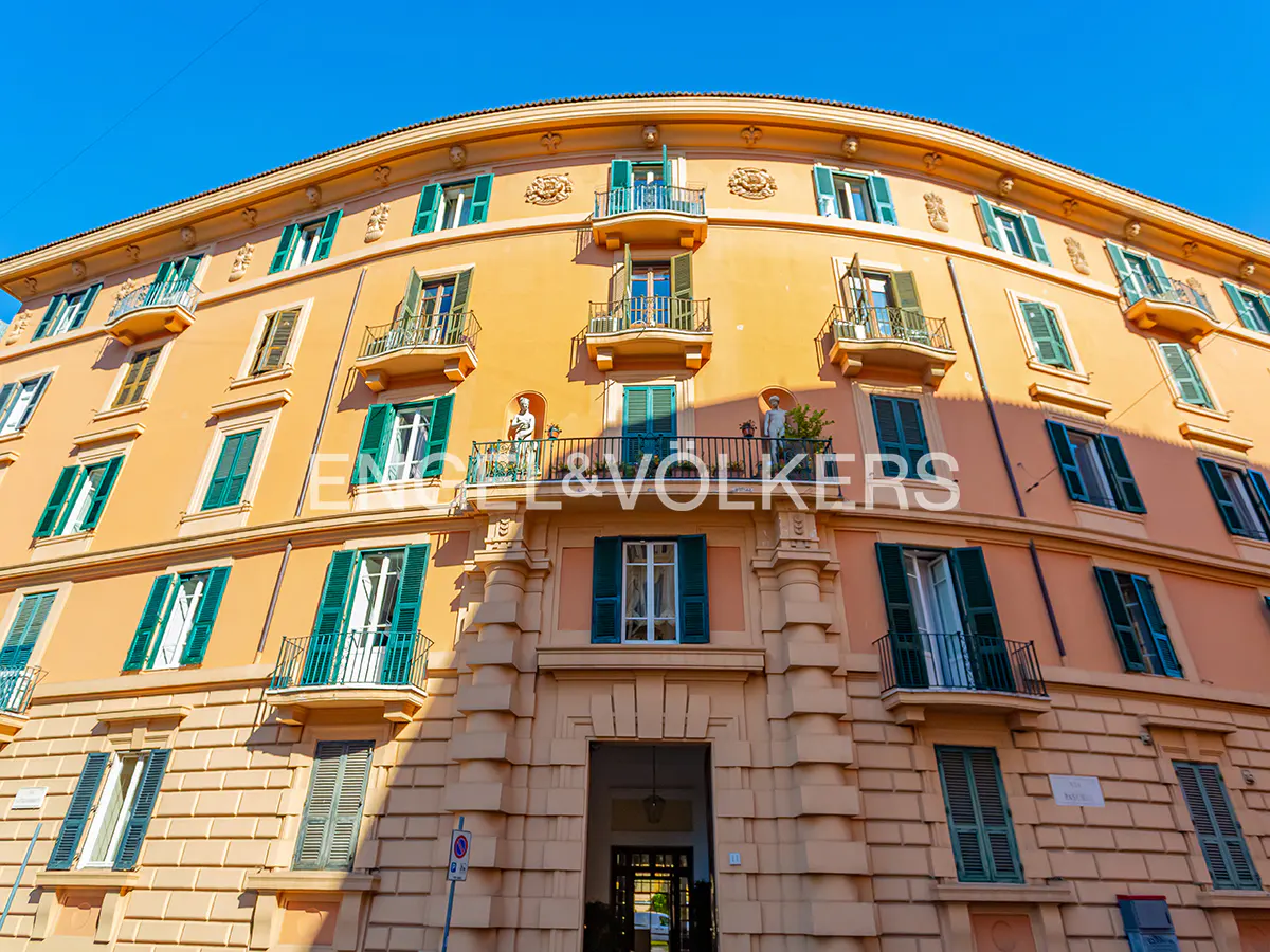 Low-angle view of a curved, peach-colored historic building with green shutters, balconies, and statues under a blue sky.