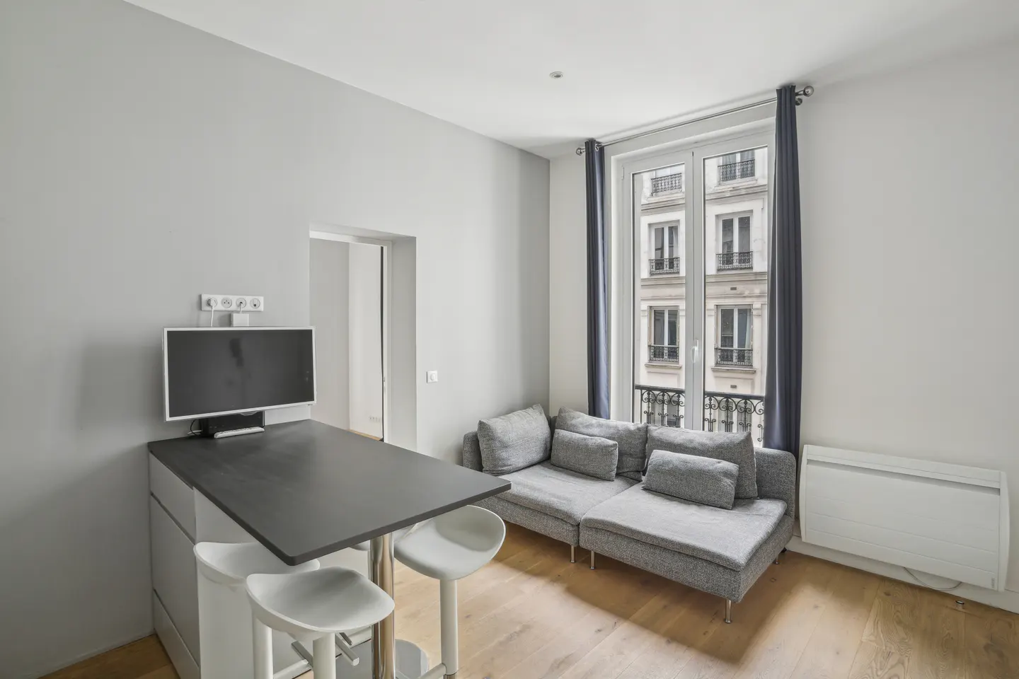 A bright studio apartment with a gray sofa, a black table with two white stools, and a window with blue curtains.