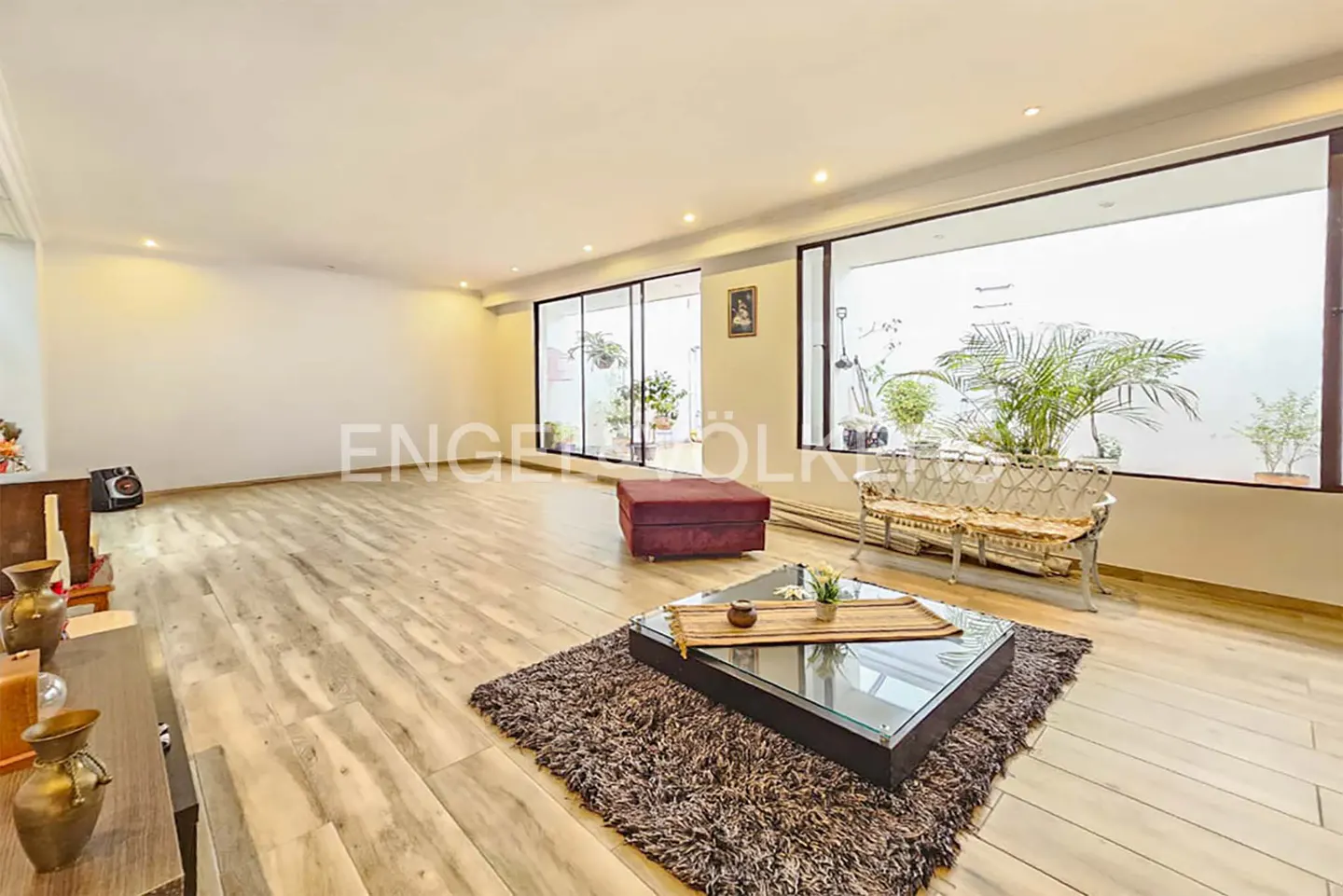 A bright living room with wood floors, a glass coffee table on a brown rug, and a red ottoman. Large windows overlook a patio with plants.