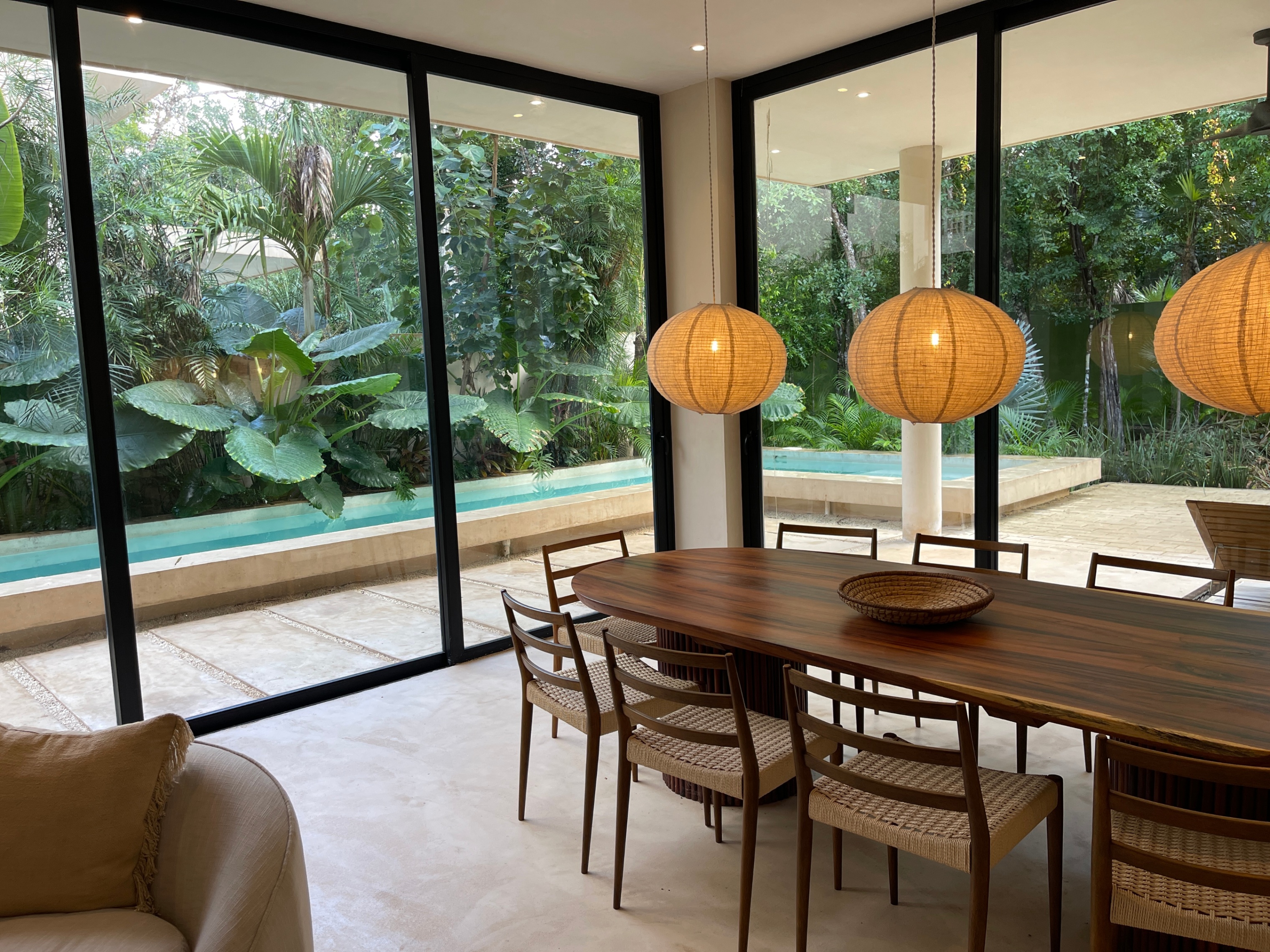 Modern dining room with a wooden table, woven chairs, and pendant lights, overlooking a lush garden and lap pool.