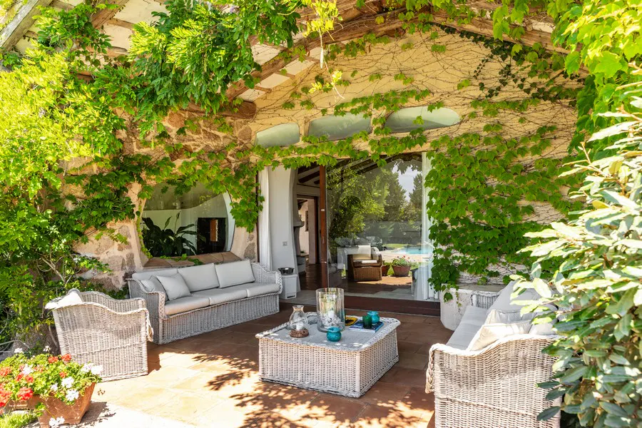 Outdoor patio with wicker furniture, stone walls, and lush green vines. Sliding glass doors lead to a pool area.