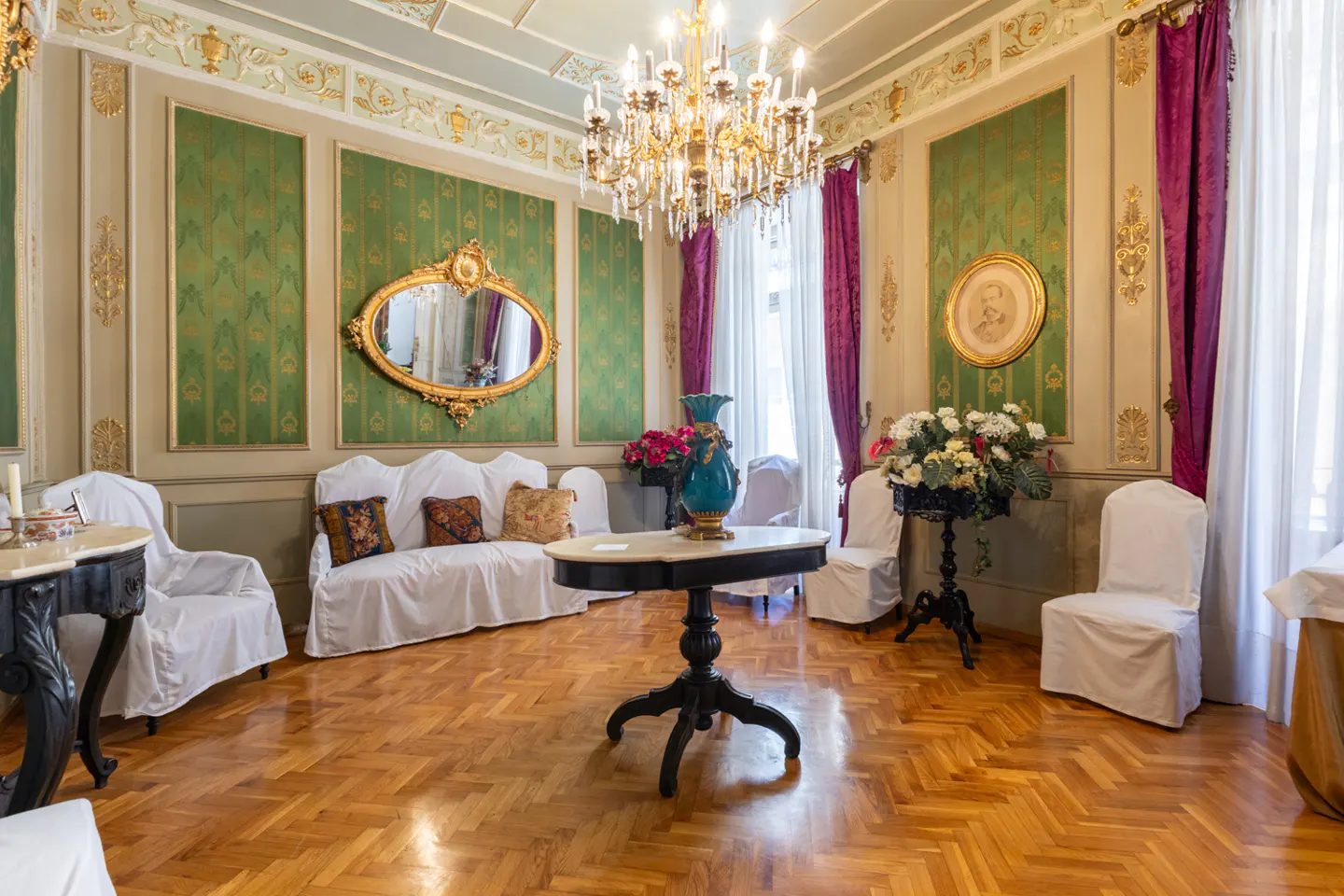 Ornate room with green patterned walls, gold trim, and a chandelier. Furniture is covered in white fabric. A round table sits in the center.