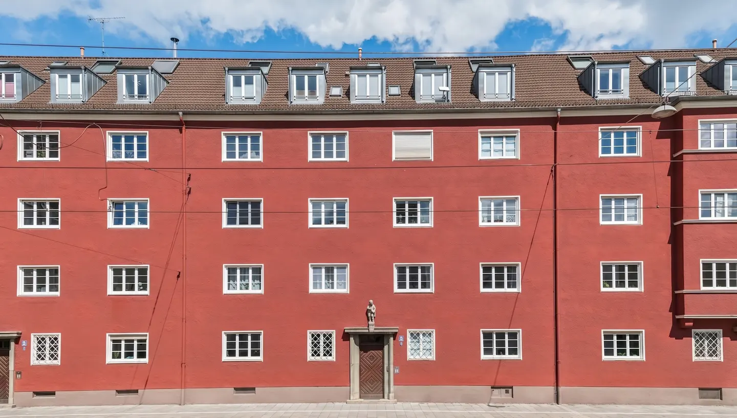 Red apartment building with white windows and a brown roof. A statue stands above the entrance door. Blue sky with clouds in the background.