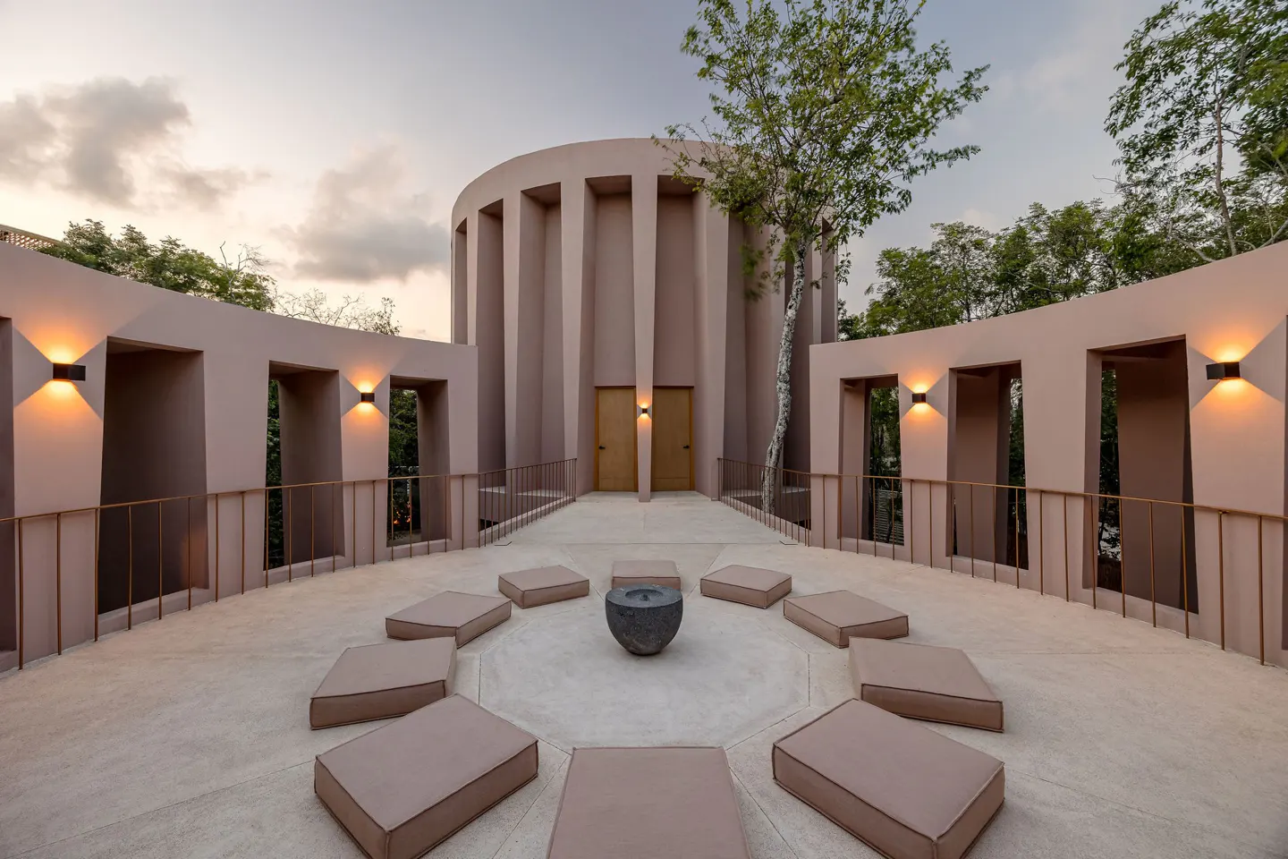 Outdoor patio with pink walls, a cylindrical building, and a circle of cushions around a dark stone bowl.
