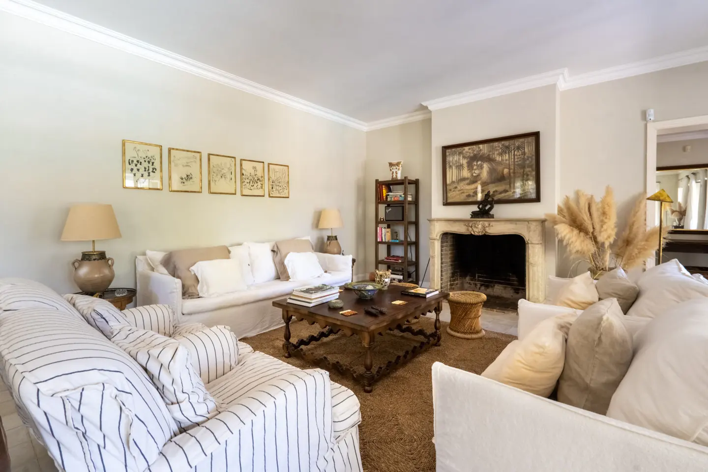 Living room with white sofas, striped armchair, and a dark wood coffee table on a brown rug. A fireplace, bookshelf, and framed art add detail.