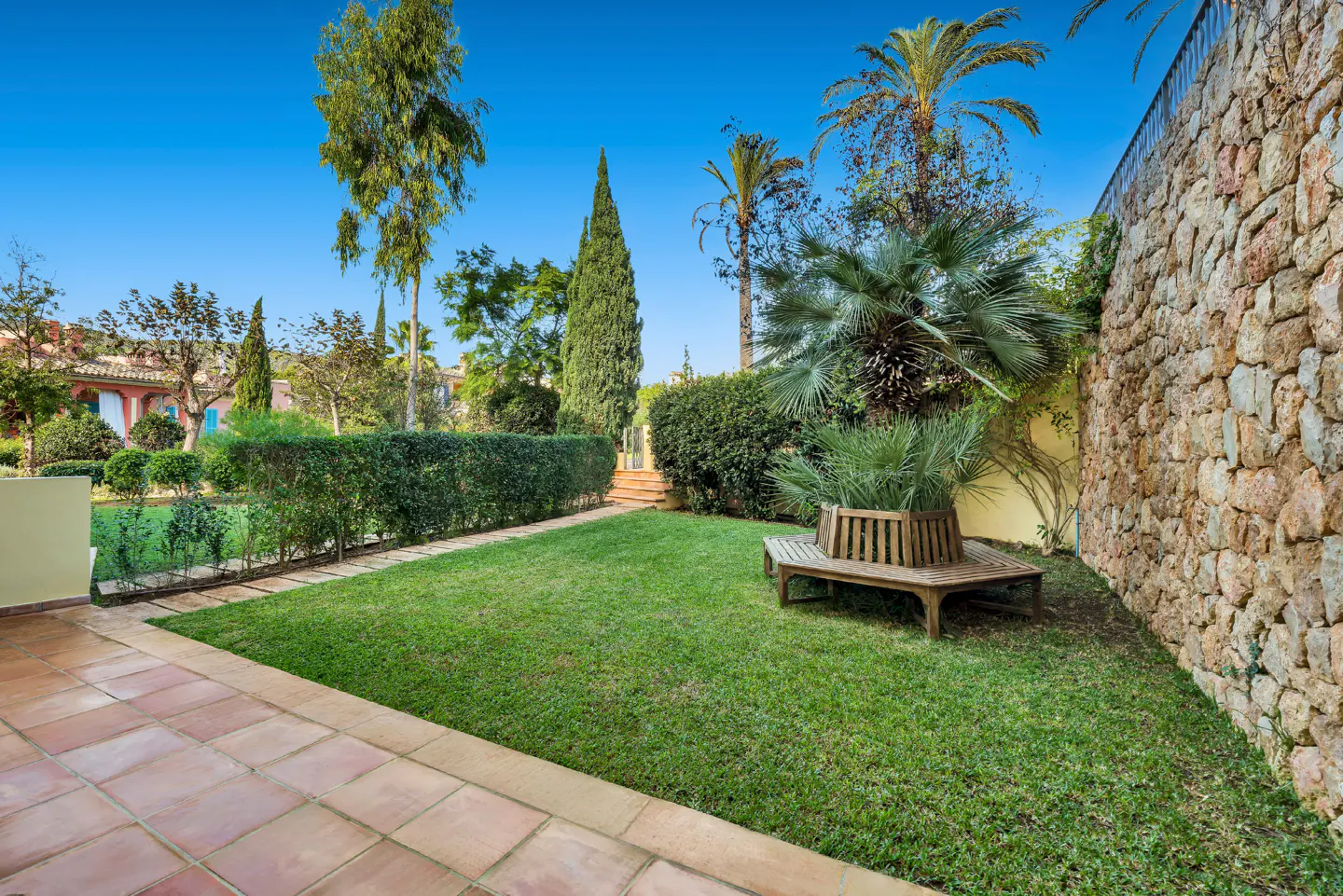 A lush green lawn with a wooden bench sits beside a stone wall and manicured hedges under a bright blue sky.