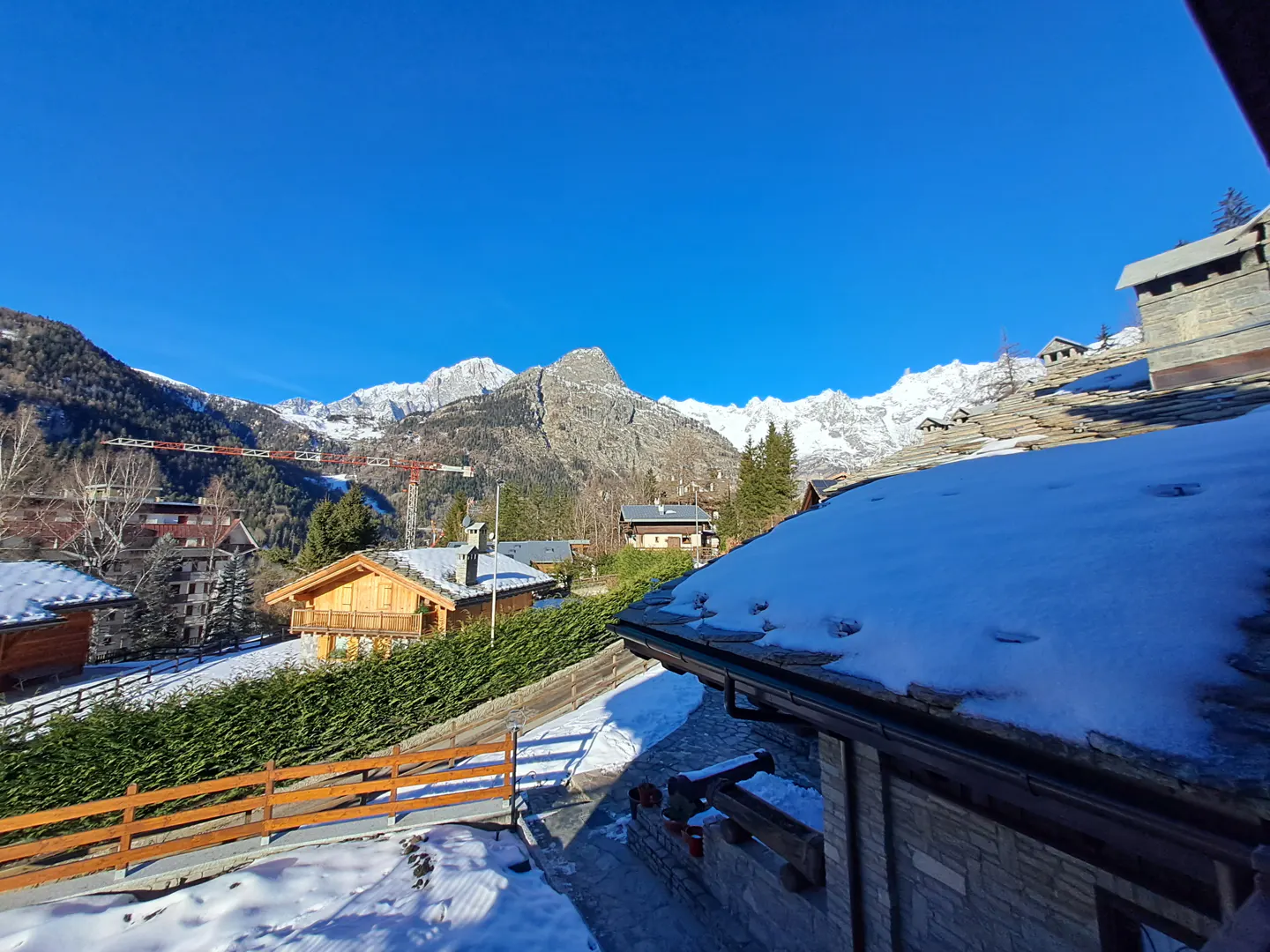 View of snow-capped mountains and chalets under a clear blue sky. A roof covered in snow is in the foreground.