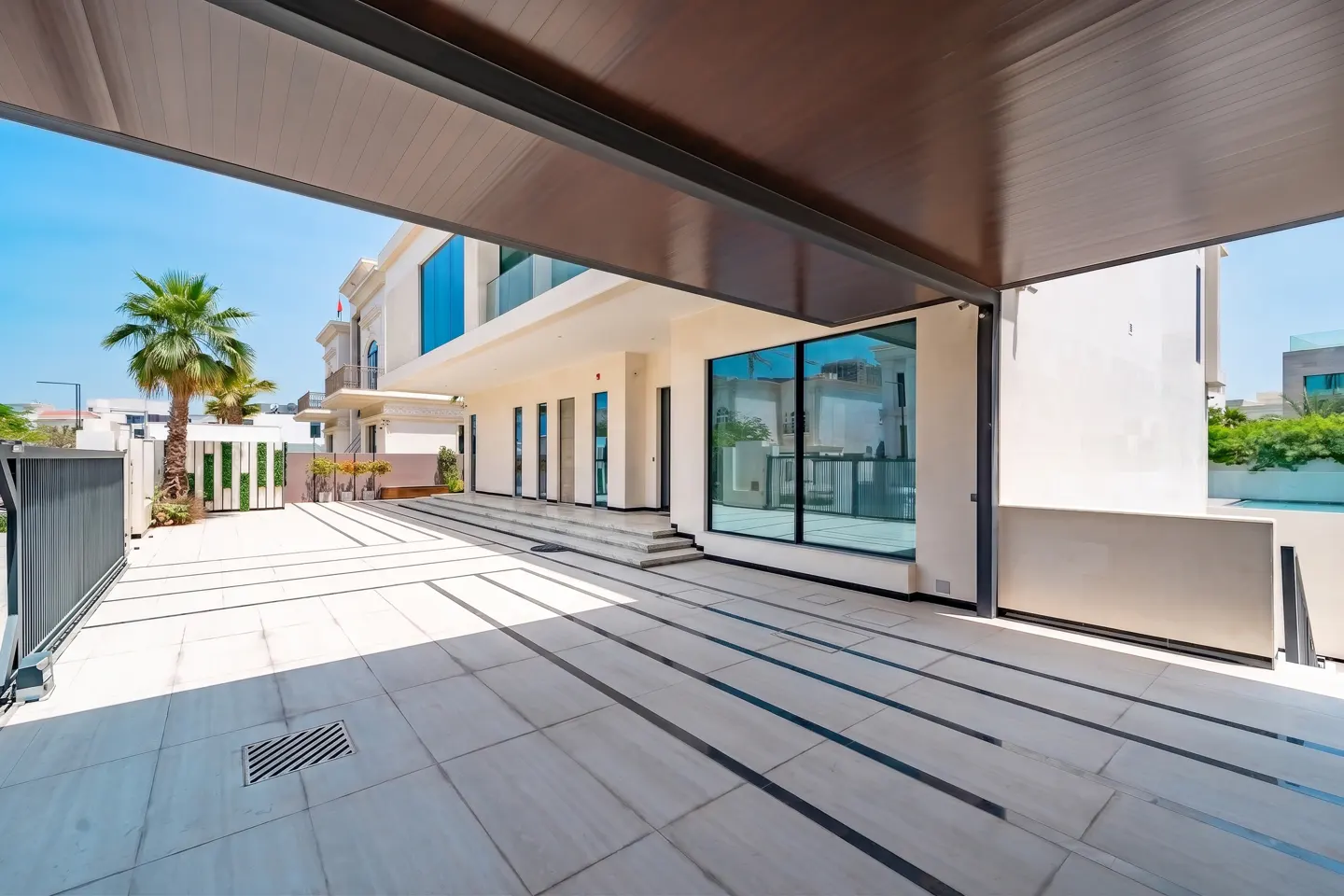 Exterior view of a modern white house with large windows and a tiled patio under a brown awning. A palm tree is visible in the background.