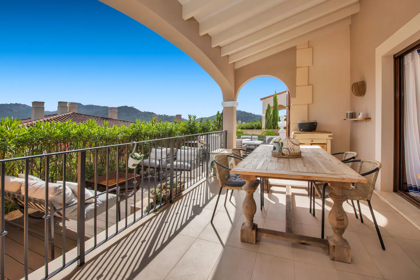 Outdoor patio with a wooden table, wicker chairs, and a lounge area overlooking a green landscape under a clear blue sky.