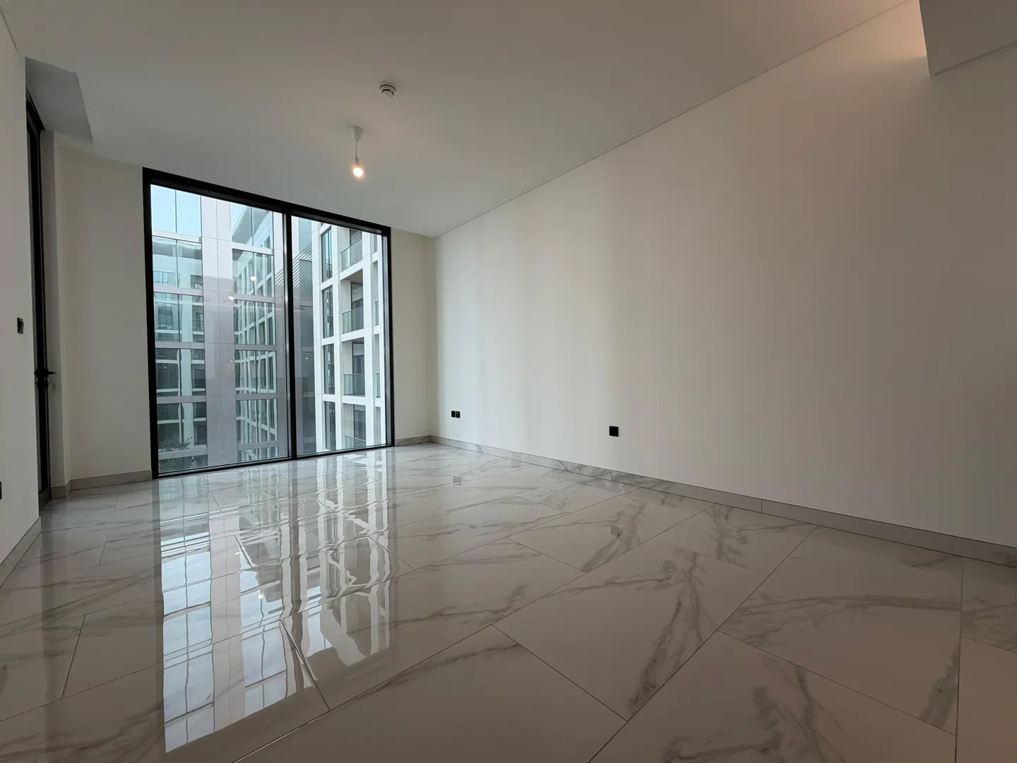 Empty room with white walls, glossy white marble-patterned tile floors, and a large window overlooking city buildings.