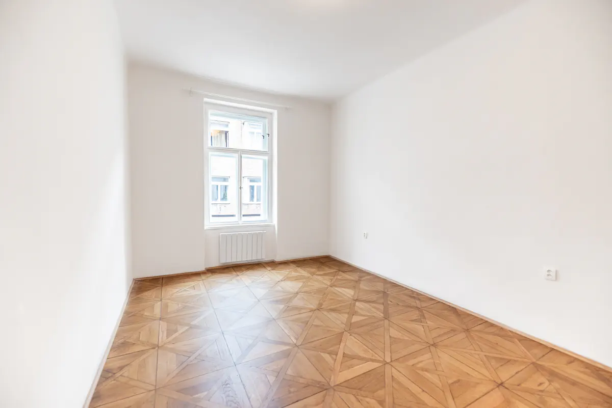 Bright, empty room with white walls, a patterned wood floor, and a window with a radiator below.