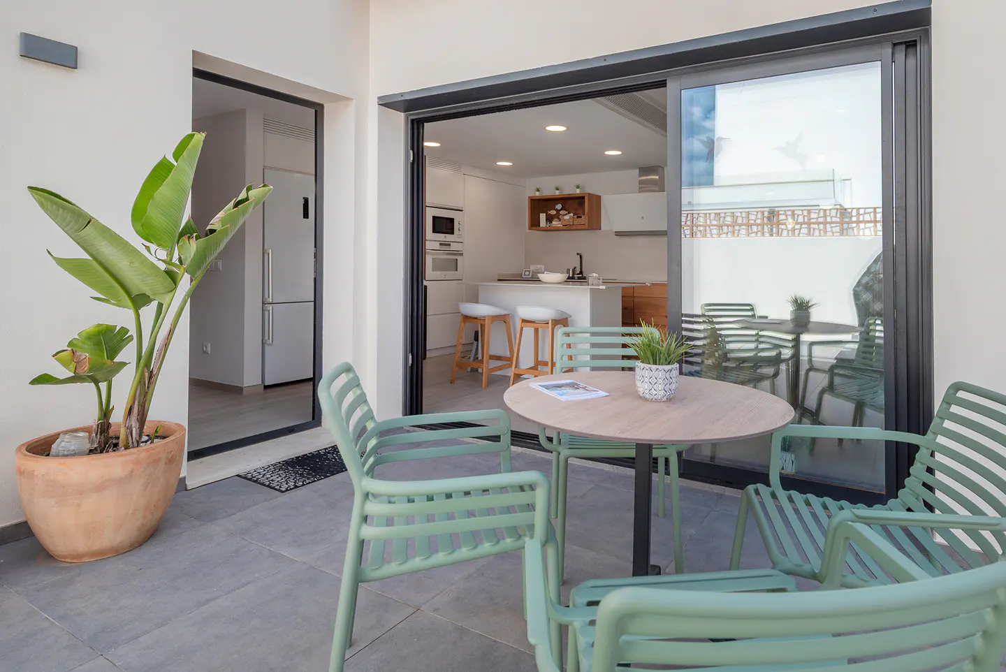 Outdoor patio with round table, green chairs, and potted plant. Open sliding doors lead to a modern kitchen with white cabinets and bar stools.