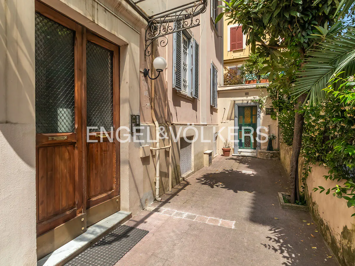A paved courtyard leads to a wooden double door and a green door, framed by a beige building and lush green foliage.