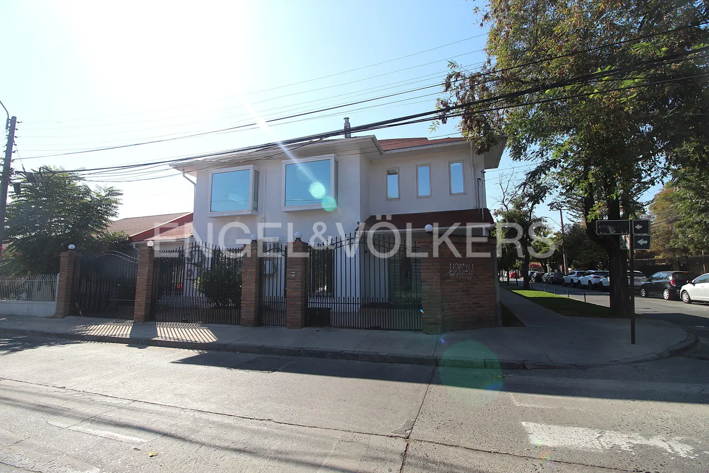 Two-story white building with the Engel & Völkers logo, black iron fence, and brick pillars on a sunny street.