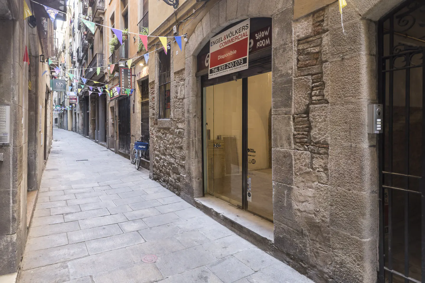 Narrow cobblestone street in Barcelona with colorful flags overhead. Engel & Volkers Commercial office with "Disponible" sign. Bike parked nearby.