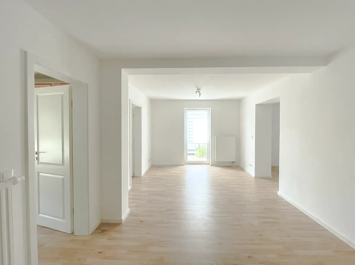 Bright, empty room with light wood floors and white walls. A door is open to the left, and a window is at the far end.