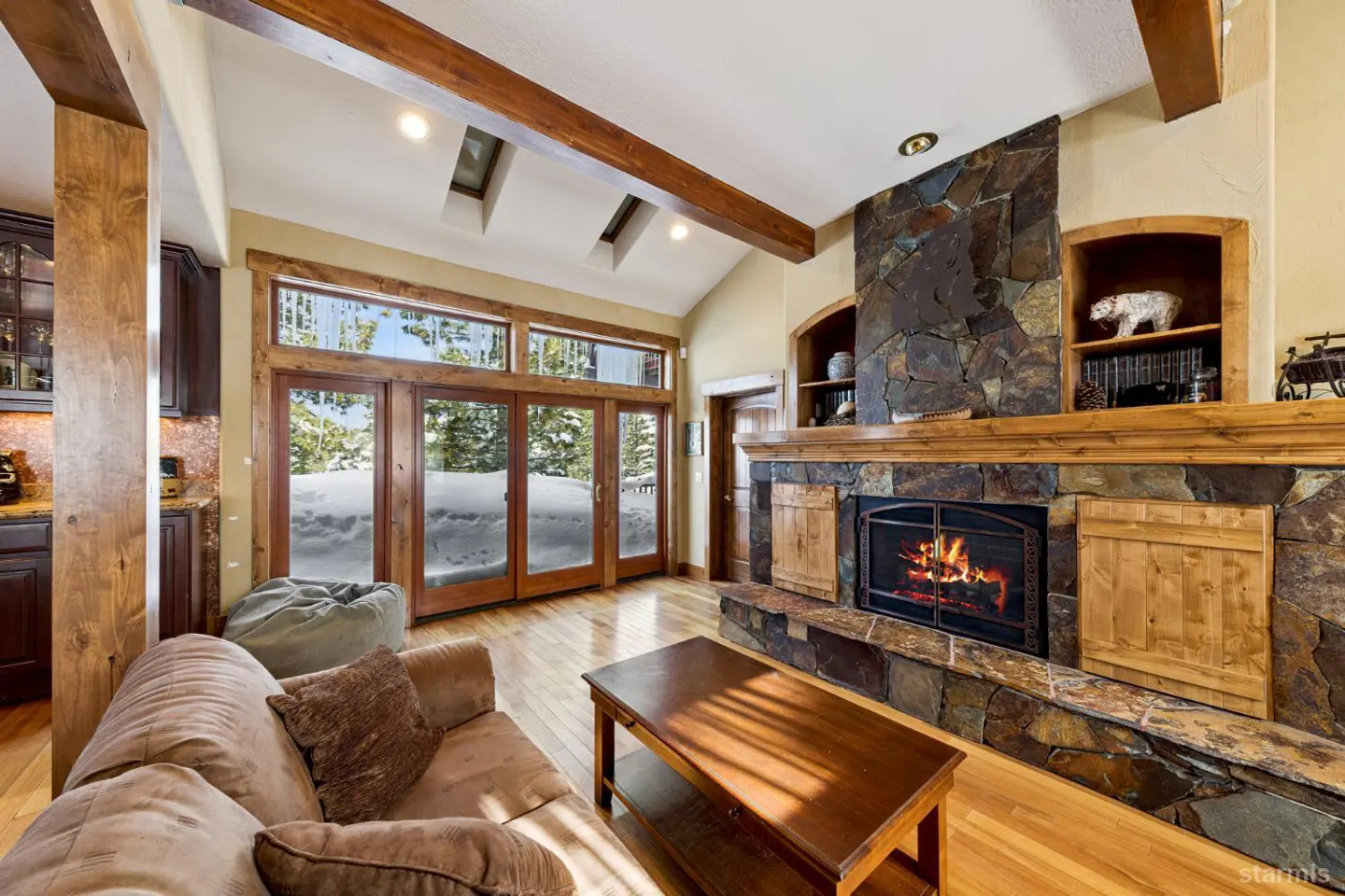 Living room with stone fireplace, wood beams, and large windows showing snow-covered landscape. Couch and coffee table in foreground.