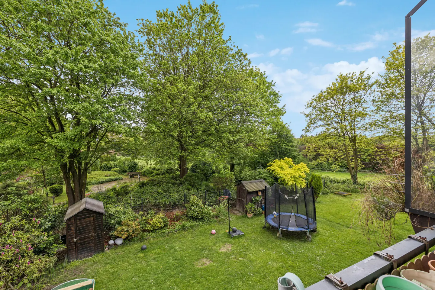 A lush green backyard with mature trees, a trampoline, shed, and garden, viewed from a balcony under a partly cloudy sky.