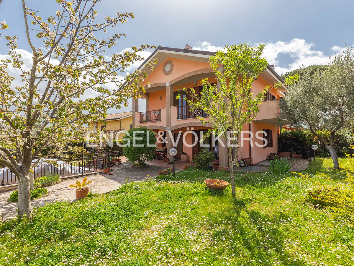 Two-story peach house with balconies, surrounded by a green lawn and trees under a sunny sky.