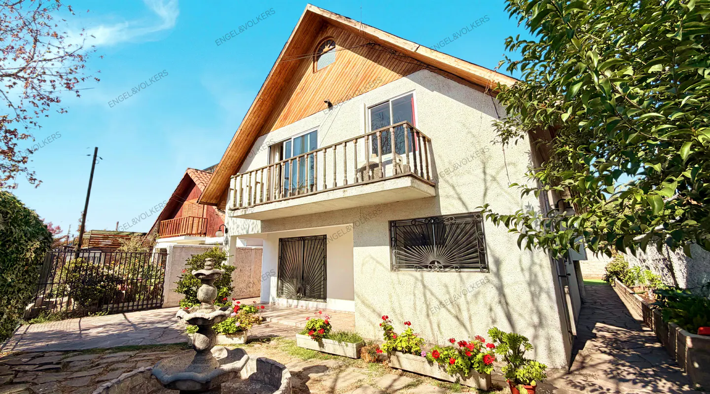 Two-story house with a wood-paneled gable roof, balcony, and a stone fountain in the front yard. Red flowers in planters.