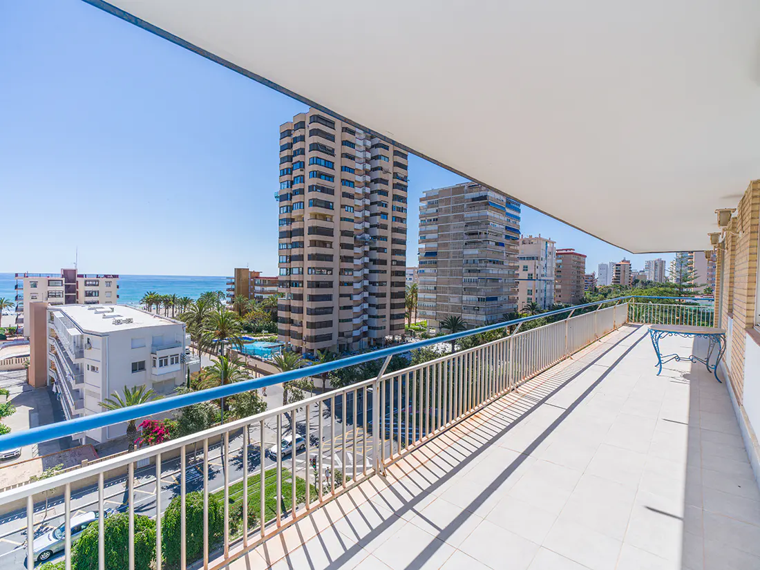 View from a balcony with white railings overlooking city buildings, palm trees, and the ocean on a sunny day.