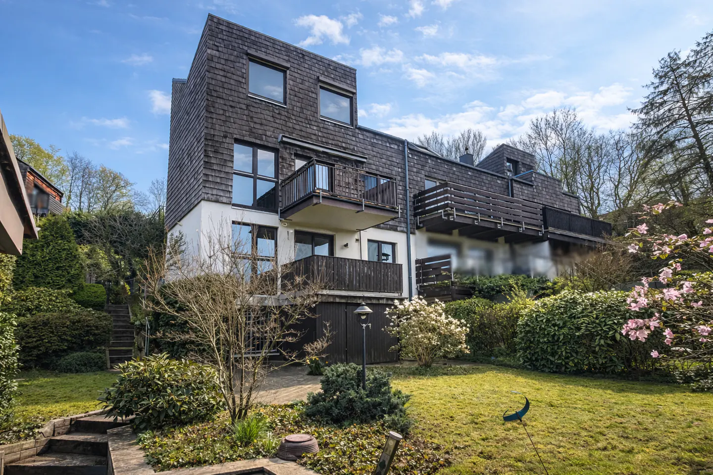 Exterior view of a modern, multi-story townhouse with dark wood shingles, white stucco, and dark wood balconies, surrounded by lush greenery.