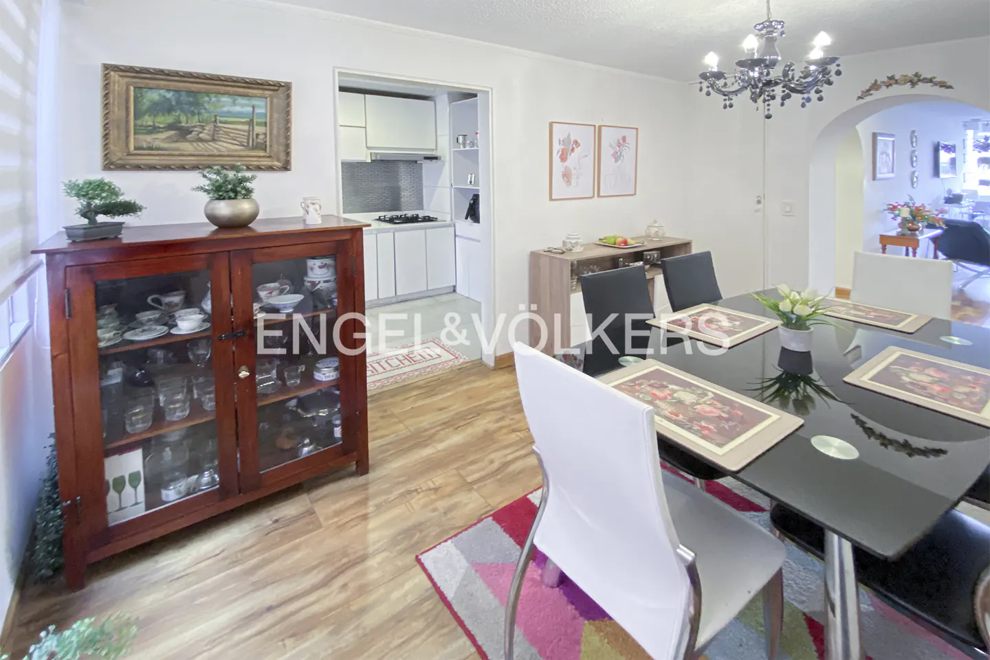 Dining room with a black glass table, white chairs, wooden hutch, and a view into a white kitchen with wood flooring.