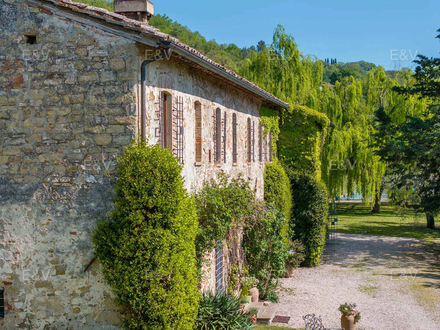 Exterior view of a stone house with green foliage, brown shutters, and a gravel path leading to a green lawn.