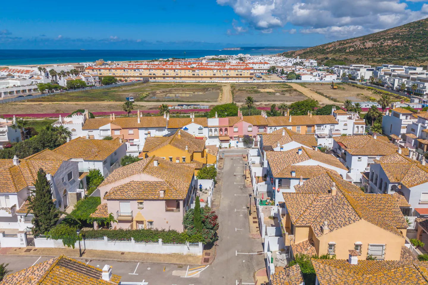 Aerial view of a coastal town with colorful houses and orange tile roofs, near a beach and green hills.