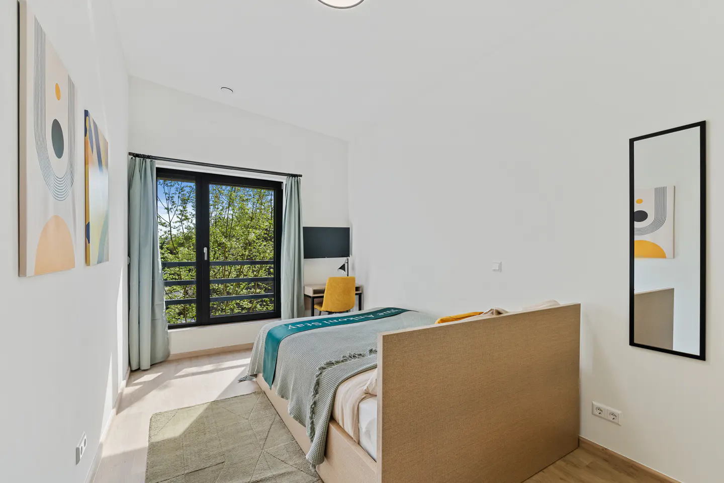 Bright bedroom with a bed, desk, and black-framed window showing green trees. Artwork hangs on the white walls, and a mirror reflects the room.